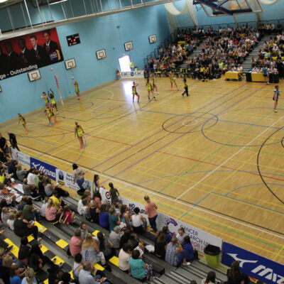 A multi sports hall with a group of young people practicing netball training. A crowd of young people sit in stands around the sports hall watching.