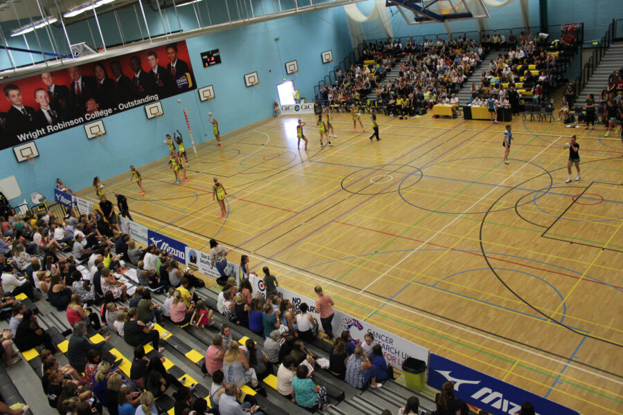 A multi sports hall with a group of young people practicing netball training. A crowd of young people sit in stands around the sports hall watching.
