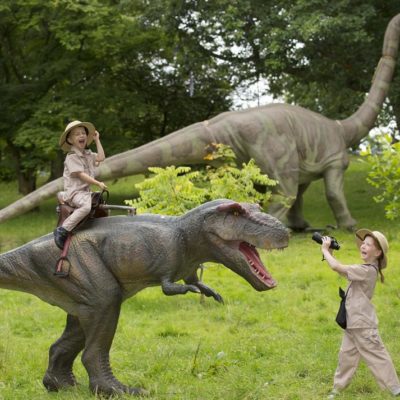 Two children dressed as explorers enjoy playing with a large model dinosaur.