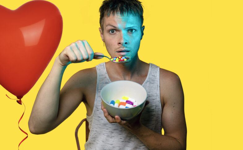 Theatre-maker Nathaniel Hall stands against a bright yellow background holding a bowl of brightly coloured pills. He hold them to his mouth on a spoon as if they are like breakfast cereal.