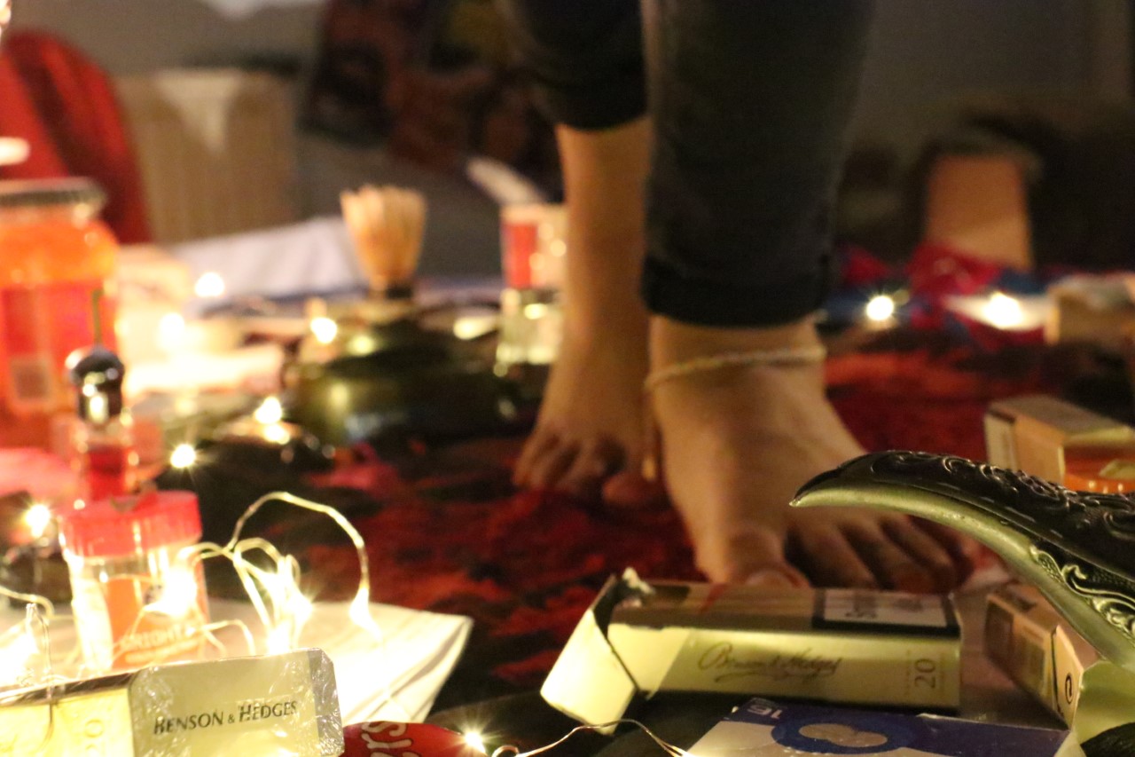 A pair of feet walk on top of a table among fairy lights and packets of cigarettes.