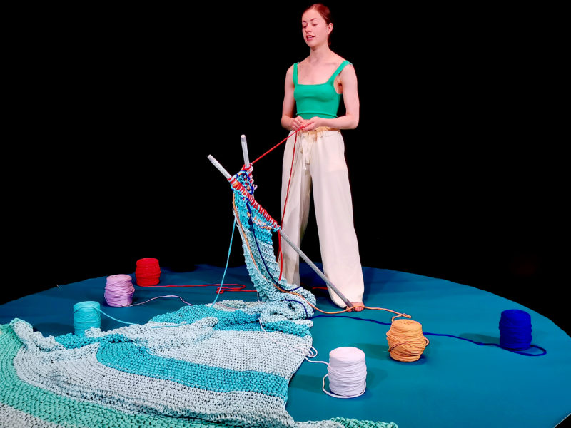 Performer Sophie Ablett stands on a stage with a huge piece of knitting, and brightly coloured balls of wool.