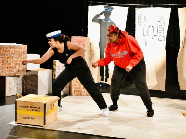Two actors perform on a stage. One wears a red hoody and the other a captain's sailor hat.