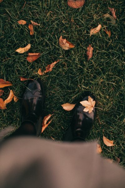 A pair of feet wearing black walking boots stands on a grassy lawn. There are autumn leaves scattered around.