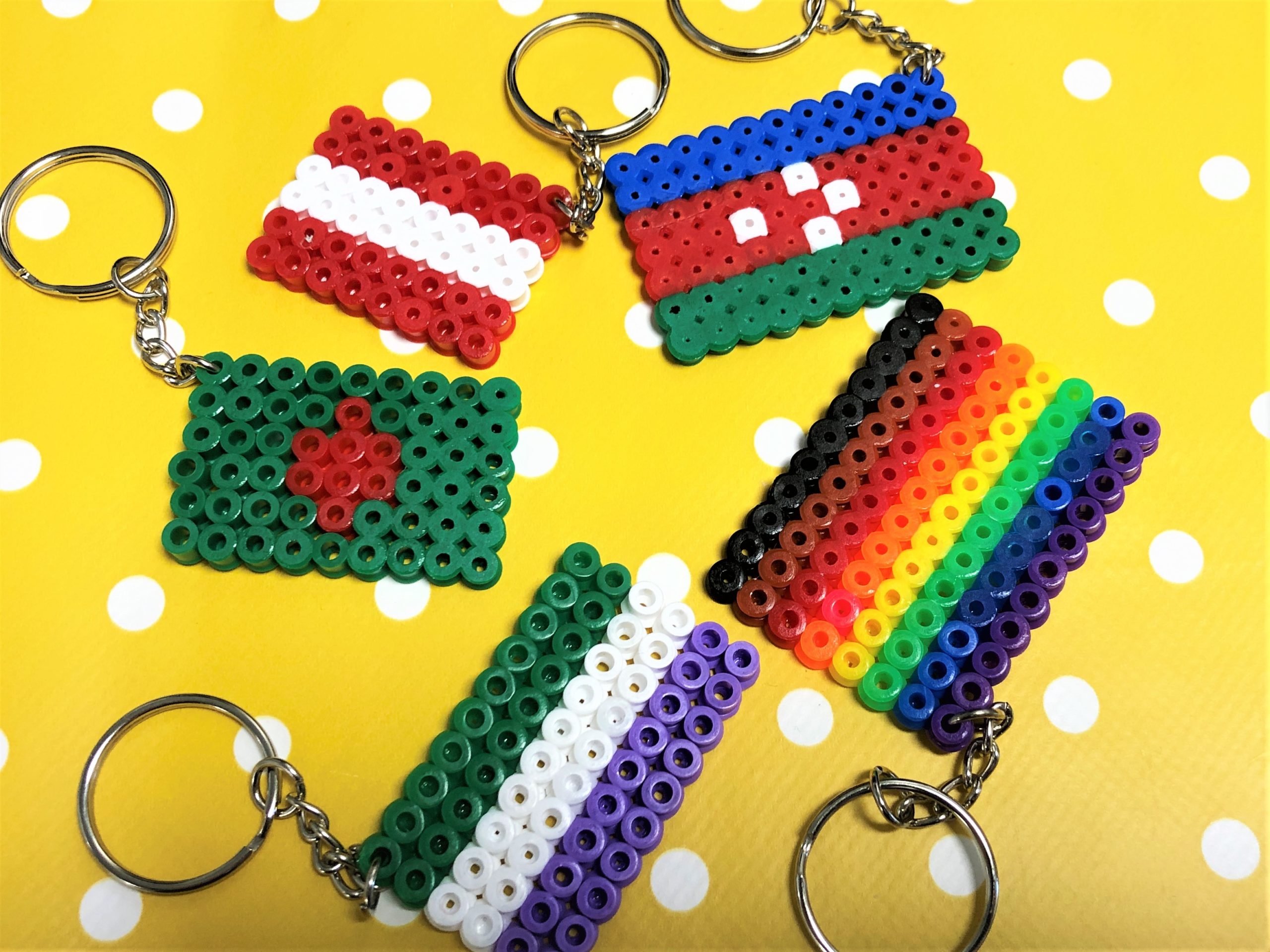 Five brightly coloured hama bead keyrings: Hama bead flag keyrings arranged in a group on a yellow and white spotty background. The flags from left to right are (1).purple, white and green horizontal stripe, (2). green with a red cross in the centre, (3).two red stripes separated by a white stripe, (4). blue, red and green stripes with four white dots in the centre of the red stripe (5). black, brown, red, orange, yellow, green, blue and purple stripe