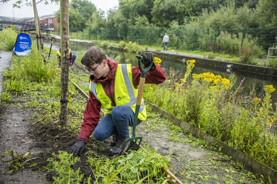 A young person wearing a high-vis kneels down to tidy a canal pathway.