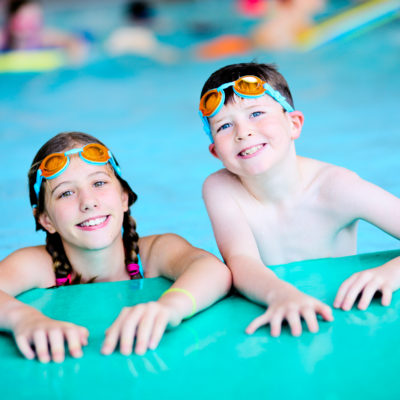 Two young people stand in a swimming pool. They are wearing goggles on their heads.