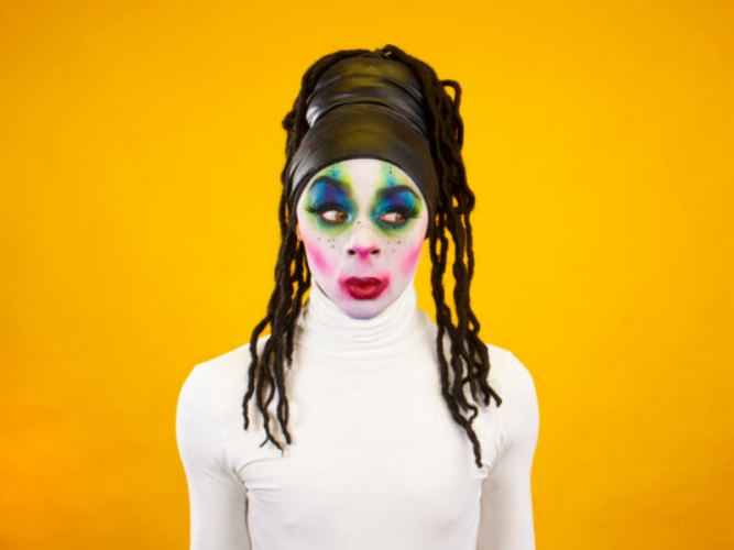 A person wearing a white turtle neck shirt, colourful makeup with a yellow background