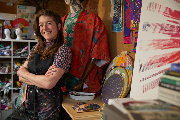 A smiling woman leans against a desk with her arms folded. she is surrounded by brightly coloured artworks that she has made in her studio