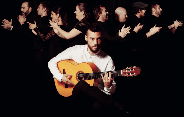 A man in a white shirt sits with an acoustic guitar, against a black background. In the background, people are stood to the side and clapping. They are dressed in black