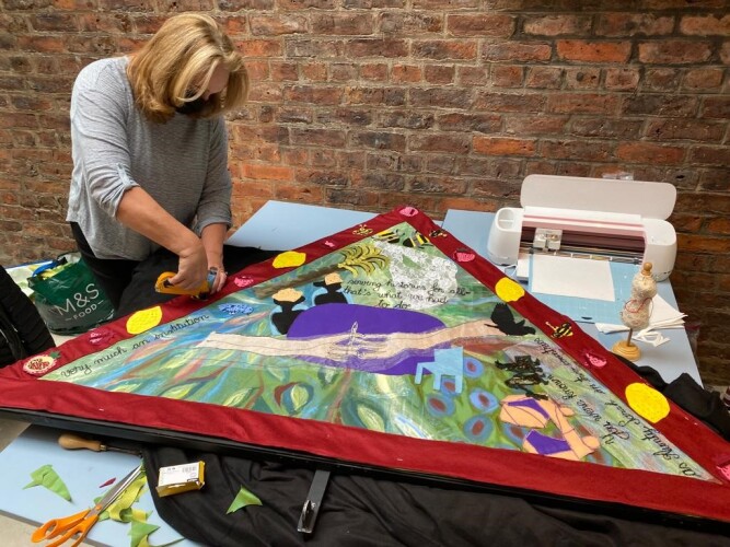 A blonde woman is holding scissors and working on a colourful textile piece.