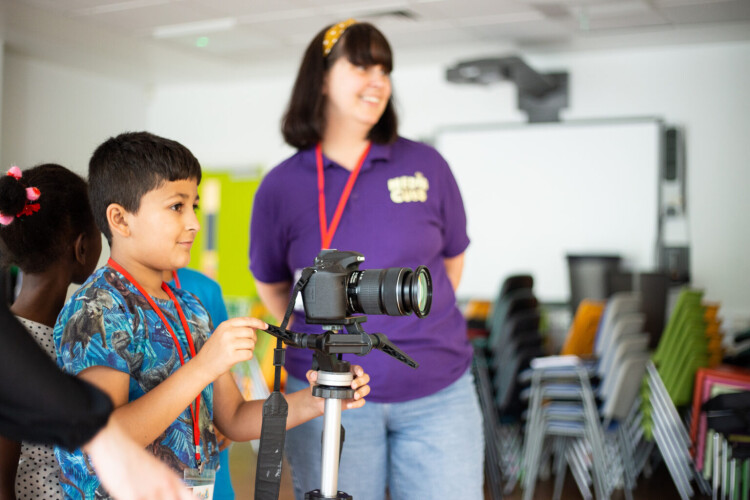 A young child using a video camera on a tripod with guidance from an adult.