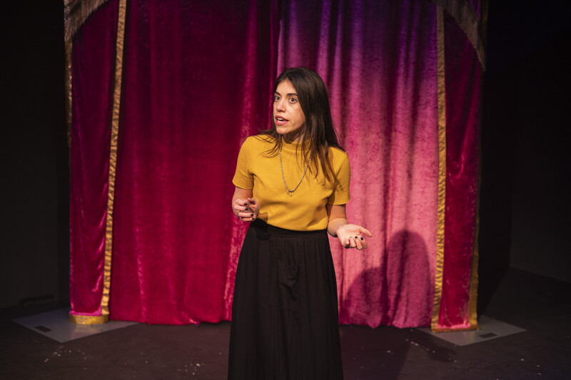 Performer Deli Segal stands on a stage infront of a red curtain.
