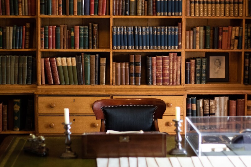 A bookshelf filled with leather bound books behind a wooden desk and chair. There are two candles in silver candle sticks on the table.