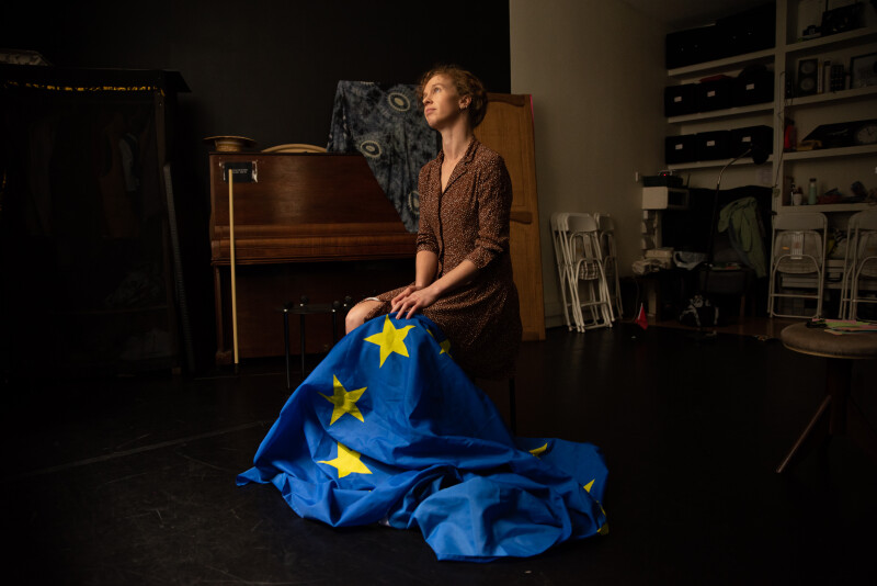 Anna Clover, a white blonde person sitting on a chair in a dark room with the European Union flag on her lap