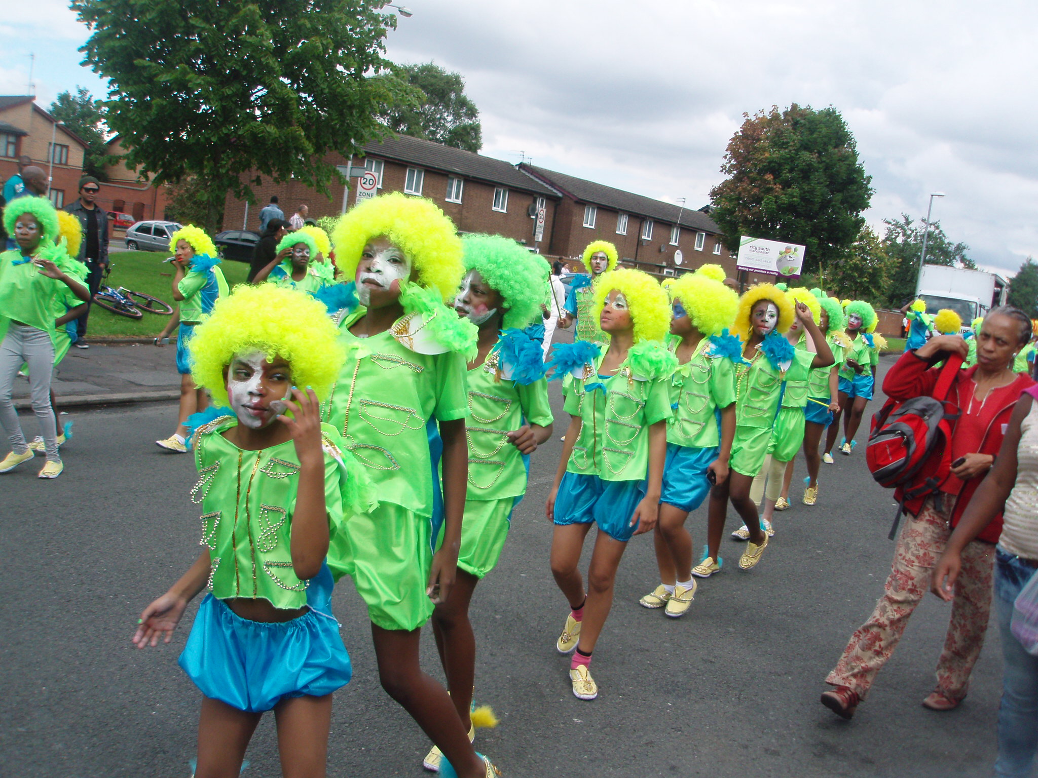 A troupe of young people dressed in green, yellow and blue walk along a road in Moss Side during the Carnival.