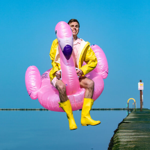 Spoken word poet Harry Baker wearing yellow wellington boots and a yellow raincoat sits on a giant inflatable flamingo as he jumps off a pier into the sea.