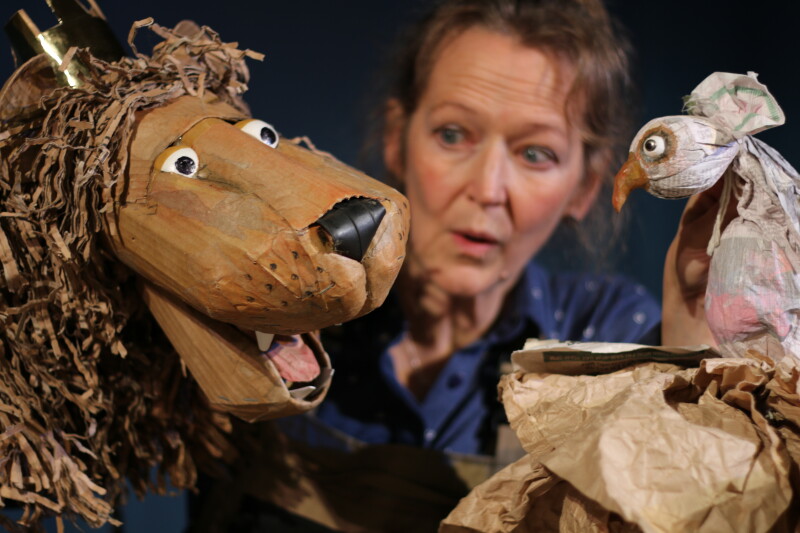 A puppeteer holds two puppets made from cardboard, a lion and a bird.