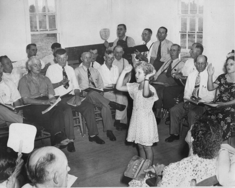 A group of people sat on chairs singing. There is a young person stood up in the middle with her arms in the air.