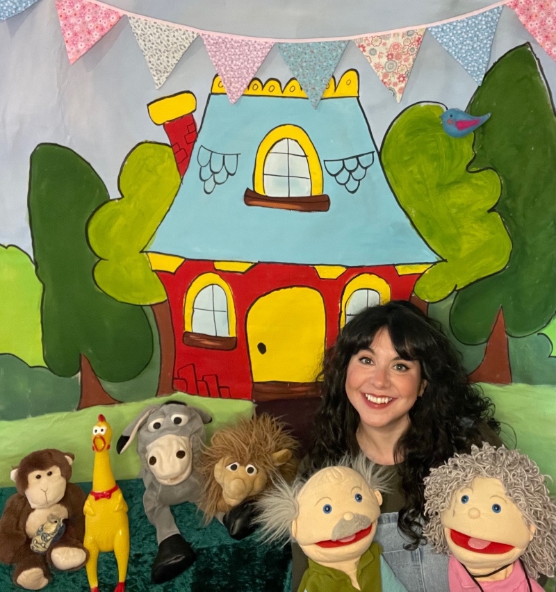 Storyteller Ilaria surrounded by her puppets and a handpainted backdrop of a house.