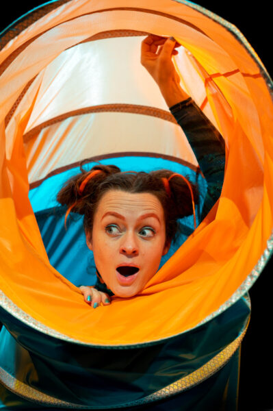 A performer emerges from a large orange tube looking surprised.