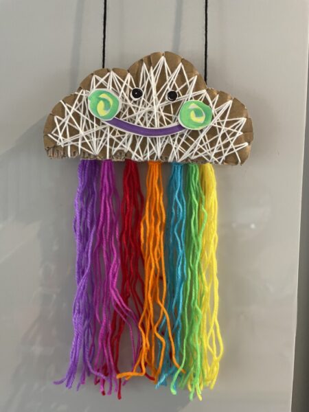 A cardboard cut-out of a cloud wrapped in wool, hanging from it is a rainbow coloured fringe.