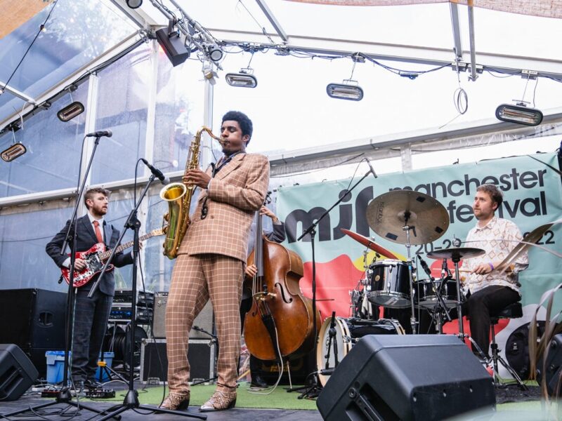 A saxophonist wearing a smart checked suit is accompanied by a guitarist and a drummer on a stage at the Manchester Jazz Festival.