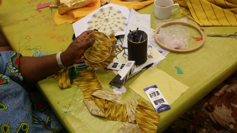 A table cloth covered in sewing materials and a person sitting down with their arm visible holding an embroidery hoop.