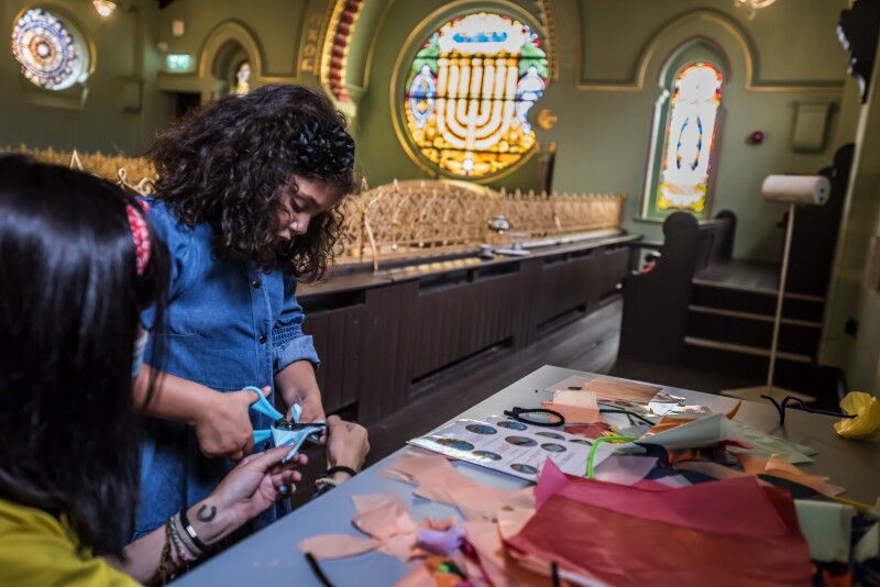 A young girl with brown, curly hair and a blue dress is cutting colourful paper with scissors, while a young woman with black hair and a yellow T-shirt assists her. They are in a synagogue, there's a round stain glass window with a menorah behind them.