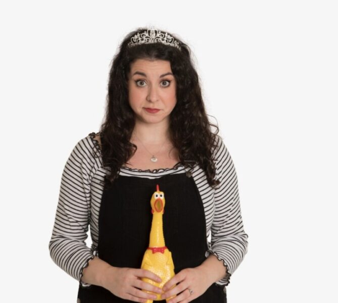 A person stands with a tiara on their head holding a plastic chicken.