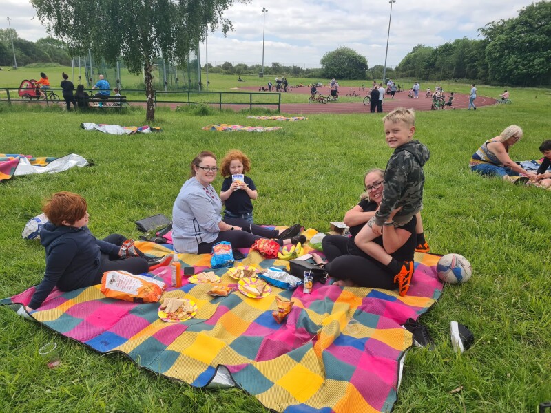 A groups of people sat on a brightly coloured picnic blanket in a field.