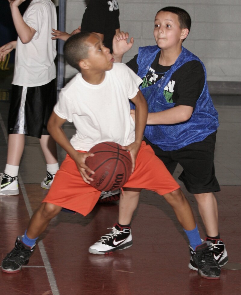 A child wearing orange shorts and a white t-shirt stands with his legs apart ready to throw a basketball.