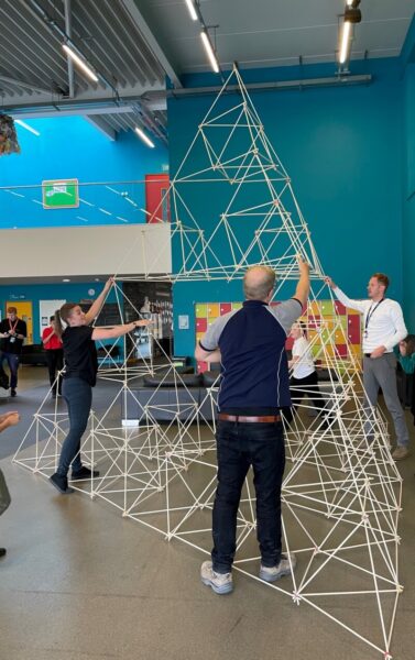 A group of people use long sticks to construct a tall geometric shape.