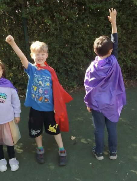 Two children dressed as super heroes and wearing capes with their arms raised in the air.