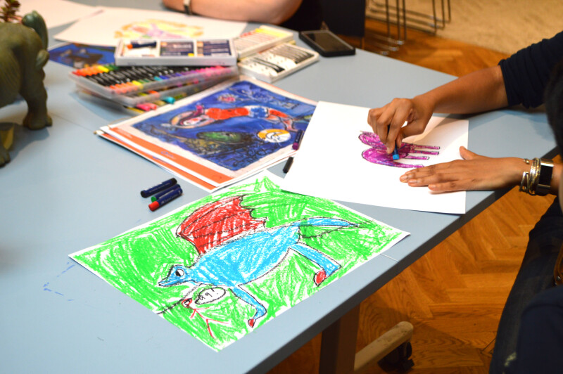 A person sits at a big grey table and draws with a blue crayon. There's another, finished drawing next to them. The drawing depicts a blue dragon with red wings on a bright green background. There is another person in the background and lots of crayons and images on the table top.