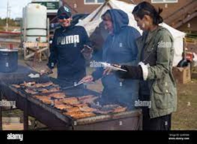 Three people stand grilling meat on a barbeque.