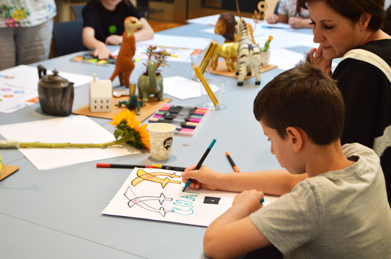 A young boy with brown hair is sitting at a big grey table and drawing with colourful pen. Next to him is an adult woman in a black T-shirt who is looking at his drawing. There are many pens, crayons and toys all over the table and other children and adults sitting in the background.