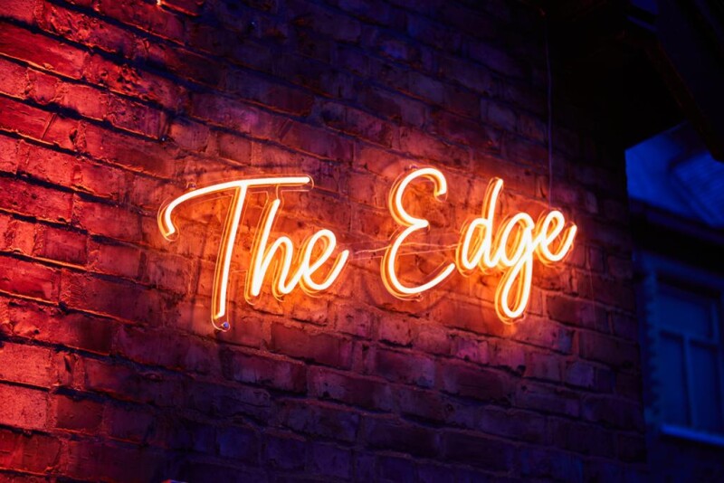 A neon sign that reads 'The Edge' against a red brick wall.
