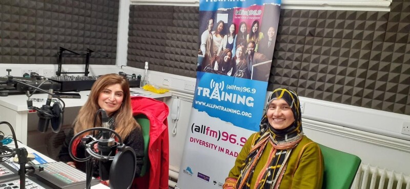 Two women smiling sat down at a table. there is a banner behind them that reads "(all fm) 96.9 Diversity in Radio".