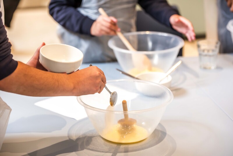 A person in a kitchen is adding ingredients to a bowl with a spoon. There is another person in the background, mixing ingredients in a bowl with a spoon.