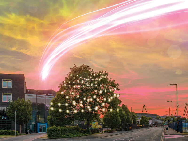 An outdoor shot of the East Manchester Academy, with the The Etihad stadium in the background. The tree in front of the academy is lit up with fairy lights and there are vibrant colours in the sky, such a fluorescent pink, orange, and yellow.