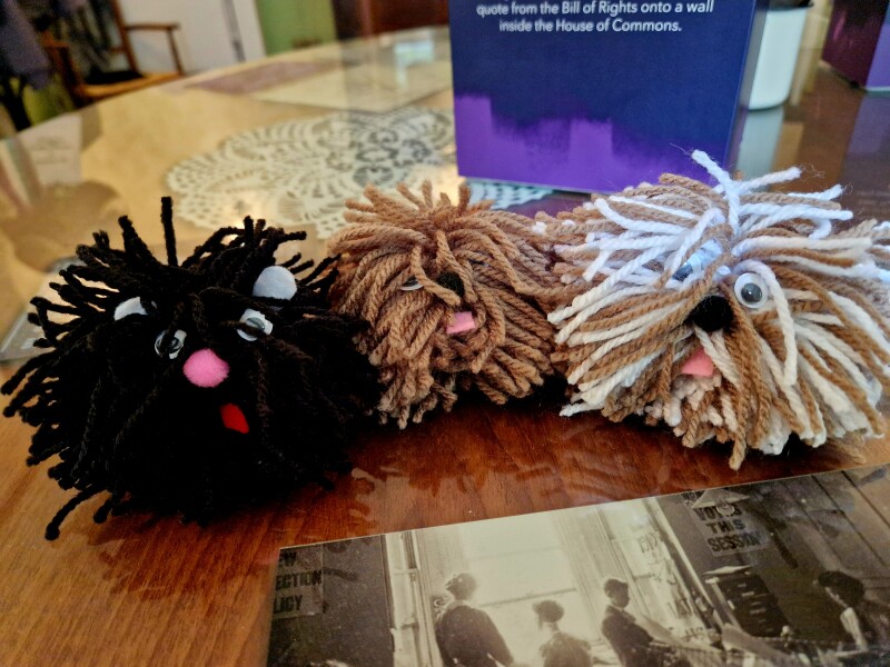 A black woollen pom pom, a brown woollen pom pom, and a brown and white woollen pom pom. All the pom poms have felt ears, plastic eyes and a felt tongue glued to them, and they are on top of a photograph on a wooden tabletop.