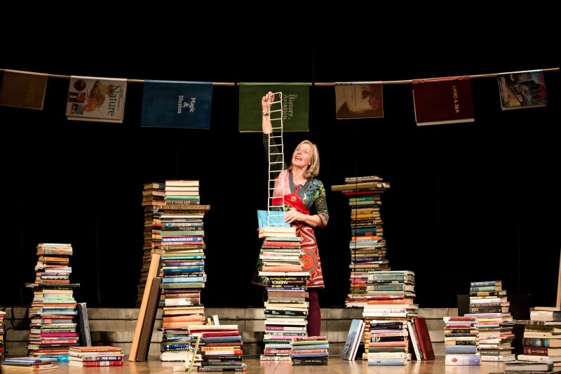 A performer stands on stage surrounded by tall piles of books.