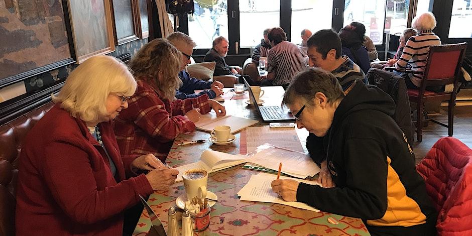 A group of older adults sit around a table doing a writing task. they have pens, paper and hot drinks.