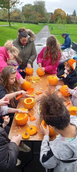 A group of adults and young people are gathered around a table carving pumpkins together.