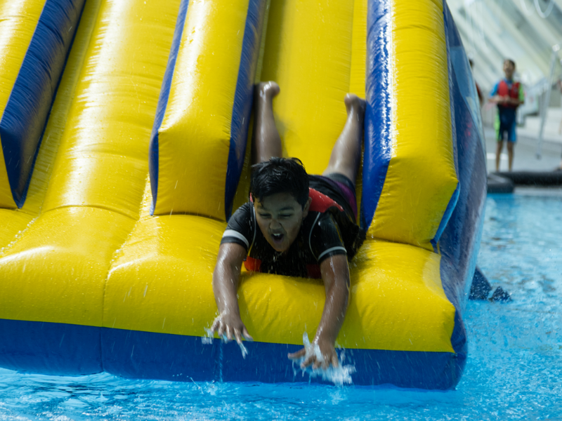A young person sliding down a giant yellow inflatable into a swimming pool.