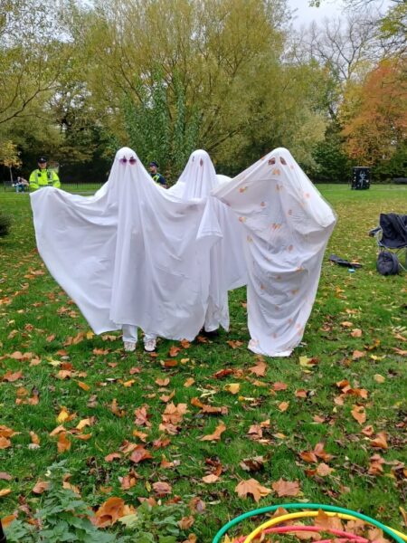 Three figures are dressed up as ghosts, wearing white sheets with their arms stretched out to the side, in a grassy field.