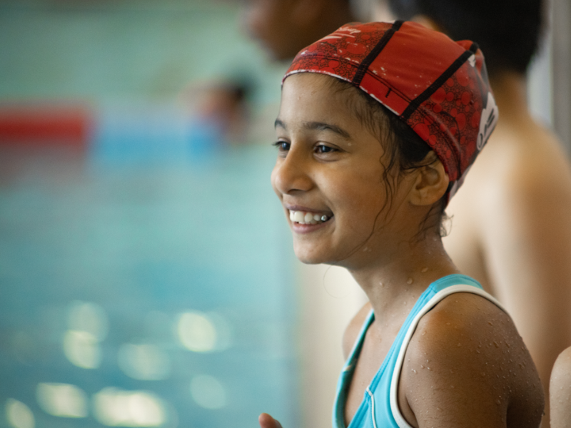 A young person with a swimming cap on stands at the side of a swimming pool smiling.