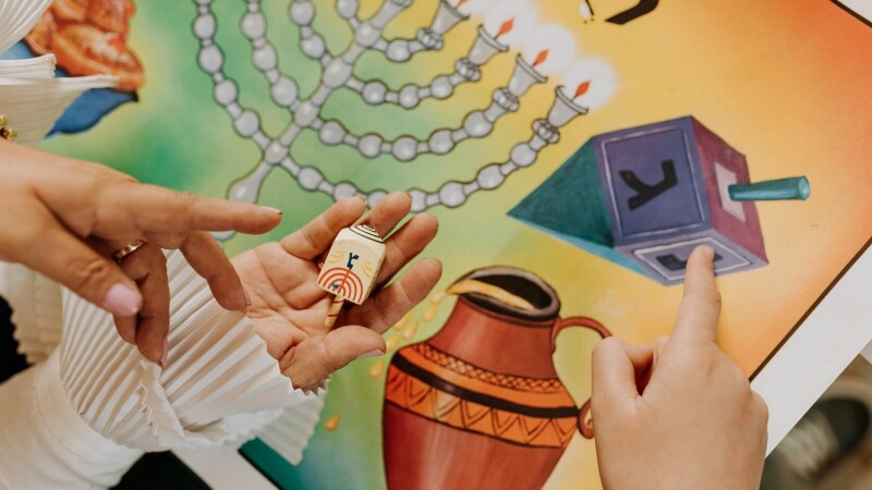 A picture of an adults and child's hands over a chanukah board game. The person on the left is holding a small wooden dreidel.