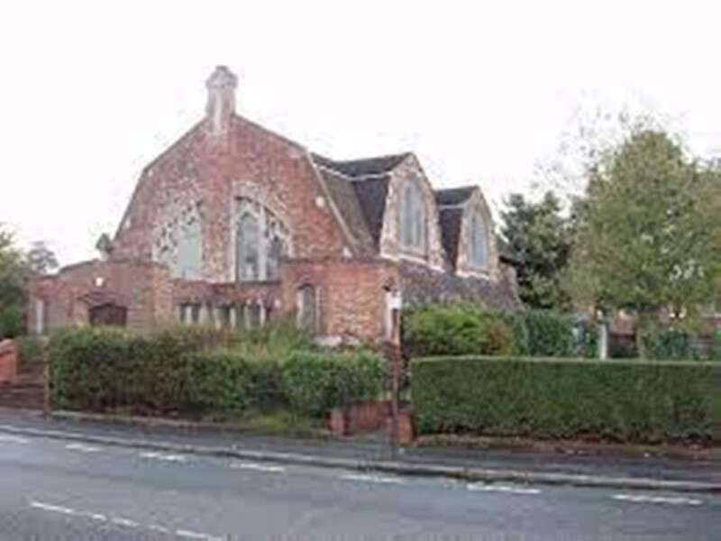 The outside of a red brick church with a hedge and trees around it.